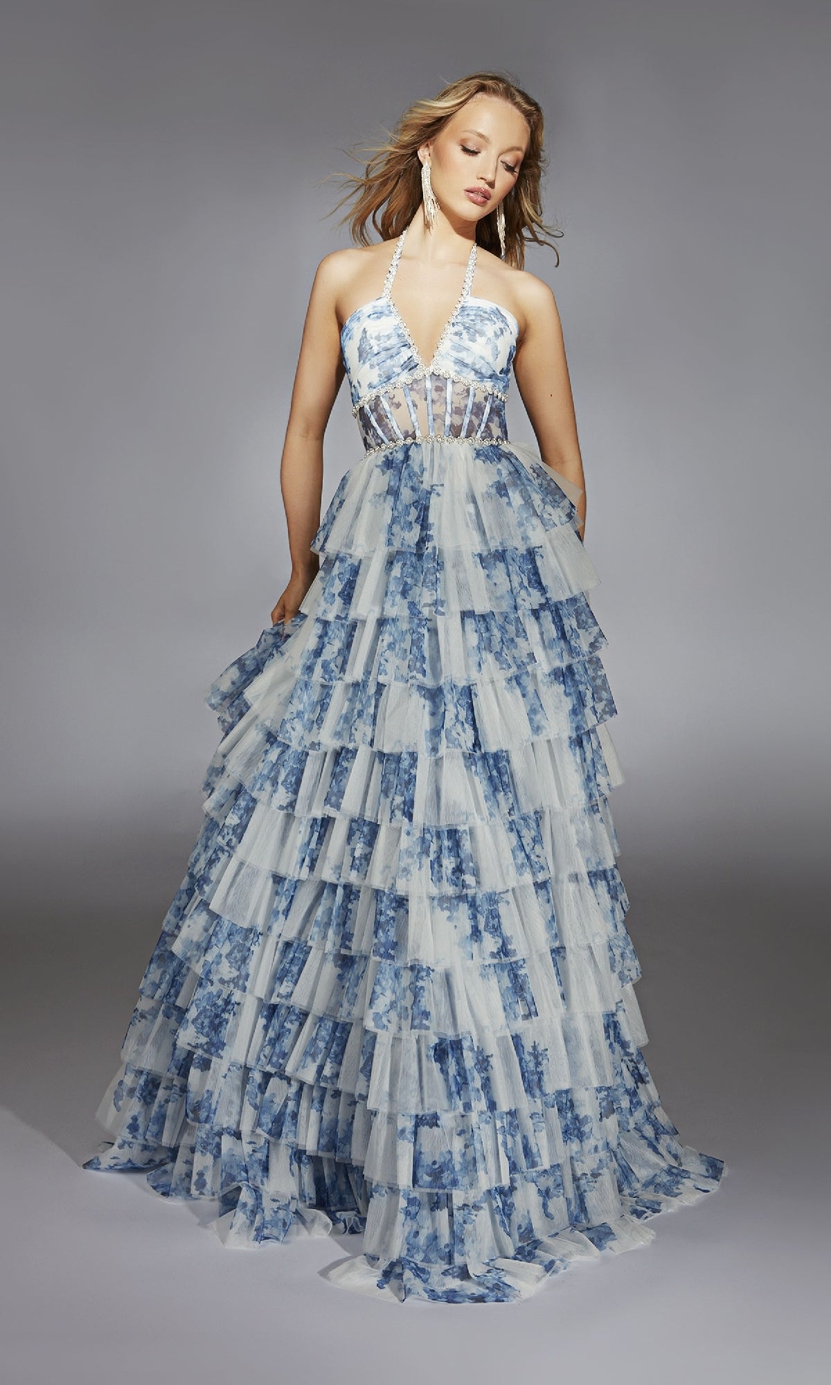 A woman in the Alyce 61730 Ruffled Long Print Prom Dress—a blue and white tiered halter gown with a sheer bodice—stands against a plain gray background.